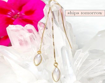 May include: A pair of gold chain earrings with a white gemstone pendant. The earrings are displayed on a white crystal background.