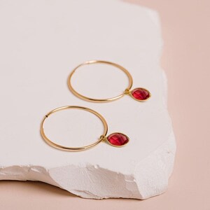 May include: A pair of gold hoop earrings, each featuring a single faceted, oval-shaped red gemstone charm suspended from the thin circular wire. The earrings are crafted from a polished gold-tone metal, showcasing a minimalist design with a secure clasp.