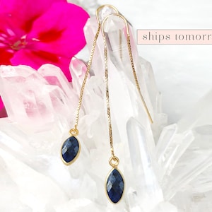 May include: A pair of gold chain earrings with a dark blue gemstone pendant on each chain. The earrings are displayed on a white crystal surface.