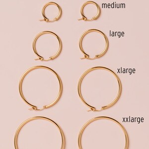 May include: A collection of gold-colored hoop earrings in various sizes, from small to xxlarge, displayed on a light pink background. The sizes are labeled with text: small, medium, large, xlarge, and xxlarge. The earrings have a classic, simple design.