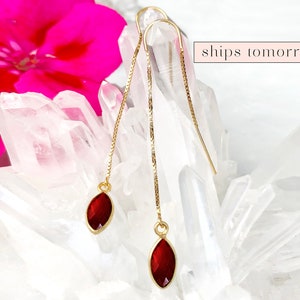 May include: A pair of gold chain earrings with red gemstone drops. The earrings are displayed on a white crystal background.