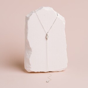 Puede incluir: Un collar tipo lariat de tono plateado con una cadena delicada y un colgante central de forma ovalada con una incrustaci&oacute;n lisa de color claro. La cadena se extiende hacia abajo desde el punto central y termina en un dije ovalado a juego.