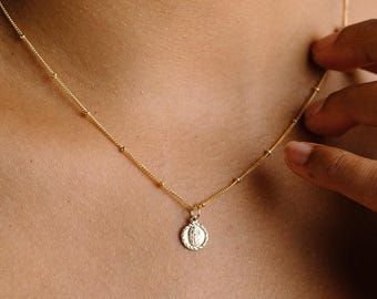 May include: A gold necklace featuring a delicate chain with small evenly spaced beads and a circular pendant. The pendant has a textured rope-style border and a central embossed figure, suspended from a simple gold jump ring.