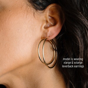 May include: Gold-colored hoop earrings. The earrings are a double hoop design, with one hoop slightly overlapping the other. The earrings are leverback style. The text in the image says "model is wearing xlarge & xxlarge leverback earrings."