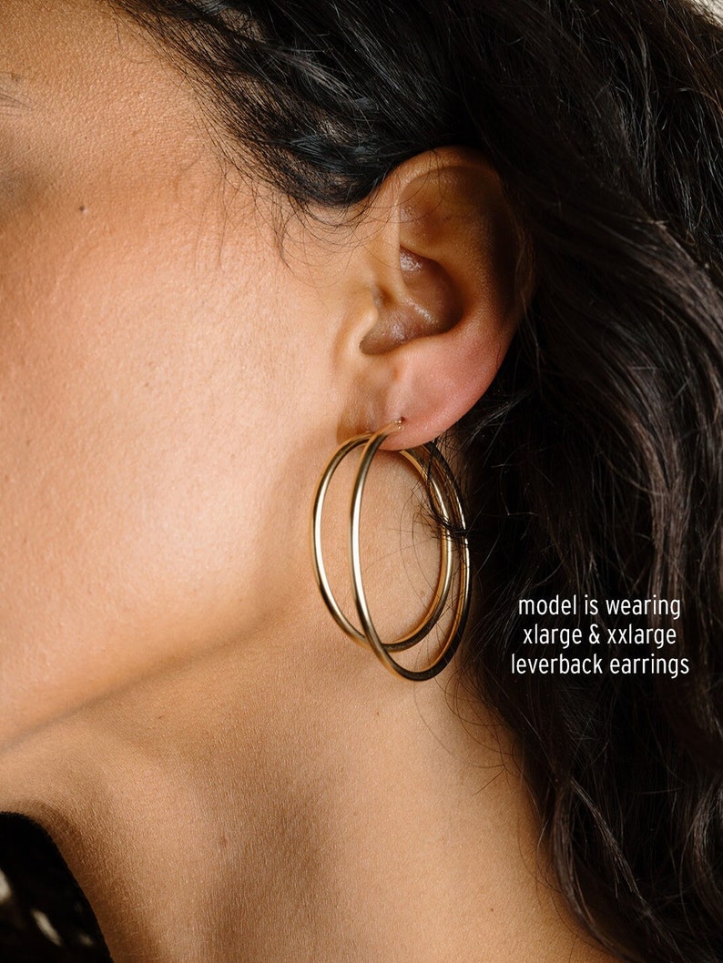 May include: Gold-colored hoop earrings. The earrings are a double hoop design, with one hoop slightly overlapping the other. The earrings are leverback style. The text in the image says "model is wearing xlarge & xxlarge leverback earrings."