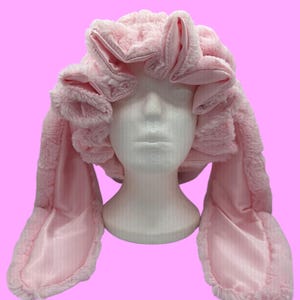 May include: A pink, fluffy bunny ear hair wrap. The wrap is made of soft, plush material with long, satin-lined ears. The hair wrap is designed to cover the head and ears, with the bunny ears extending outwards.