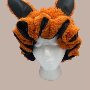May include: A plush orange and black fox ear headband. The headband features fluffy orange faux fur with black satin accents and upright ears. The ears are lined with black satin. The headband is displayed on a white mannequin head.