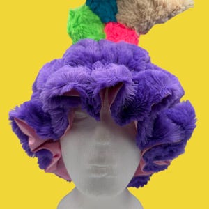 May include: A whimsical, fluffy hat in vibrant colors. The hat features a large, purple, faux fur base with a pink satin lining. A cluster of colorful faux fur accents in green, teal, pink, and tan adorns the top, creating a playful and unique design.