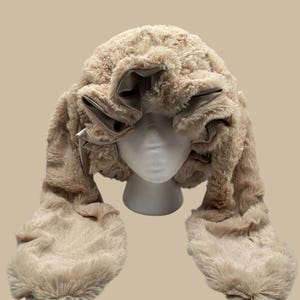 Peut inclure: Un chapeau en fausse fourrure beige avec des rabats d'oreilles attachés. Le chapeau est présenté sur une tête de mannequin blanche. Le chapeau a une texture douce et moelleuse et une couleur neutre, adaptée au froid. Les rabats d'oreilles s'étendent vers le bas, offrant une chaleur supplémentaire.