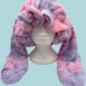 May include: A plush hat made of soft, faux fur in a tie-dye pattern of pink and lavender. The hat features a wide, ruffled brim and two long, dangling side panels that extend downwards, displayed on a plain white mannequin head.