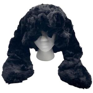May include: A black faux fur hat with long ear flaps. The hat is displayed on a white mannequin head. The fur appears soft and plush, with a slightly textured surface. The hat's design includes a rounded crown and extended ear coverings.