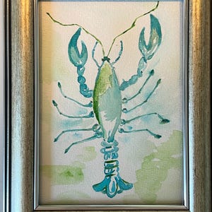 May include: Watercolor painting of a lobster in shades of teal and green, framed in a silver-toned wooden frame. The lobster is depicted with detailed claws, legs, and antennae. The background features soft green and white watercolor washes.