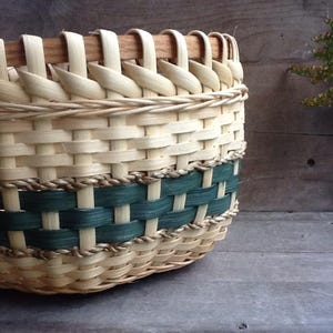 May include: A woven basket with a natural, light-coloured weave and a dark green band. The rim features a wooden handle. The basket is made of woven strips of wood and has a sturdy, rustic appearance. The basket is likely used for storage or decoration.