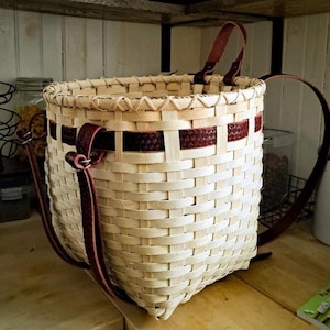 Può includere: Un cesto intrecciato a mano con una trama in legno chiaro naturale. Presenta una fascia di materiale intrecciato rosso scuro al centro e cinghie in pelle marrone scuro. Il cesto è progettato con una parte superiore arrotondata e una base quadrata.