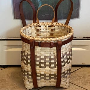 Puede incluir: Una mochila tipo cesta tejida con material de mimbre de color claro y bandas horizontales en tonos marrones y verdes apagados. La cesta incluye dos correas de hombro de cuero marrón, un asa superior y un cinturón de cuero con hebilla metálica.