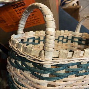 May include: A handwoven basket with a sturdy handle. The basket features a natural, light-colored weave with accents of teal green. The handle is wrapped with the same natural material, providing a comfortable grip. The basket is designed for carrying items.