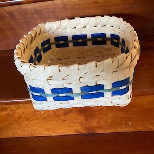 May include: A square woven basket with a natural, light-colored base and blue accents. The basket is made of woven strips of material, with blue bands and a green band adding color and detail. The basket is sitting on a wooden surface.