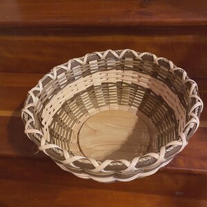 May include: A handwoven basket with a round shape and a light brown wooden base. The basket's sides are woven with alternating light and dark brown strips, and the rim features a decorative woven pattern.