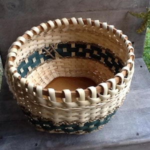 May include: A round woven basket featuring light beige natural splints and dark green accent bands. The basket has a sturdy wooden base and a thick, looped rim design. The weave pattern alternates between horizontal and vertical strips.