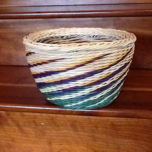 May include: A woven basket with a rounded shape and a wide opening. The basket features a spiral design with bands of white, green, yellow, purple, and brown. The basket is sitting on a wooden surface.