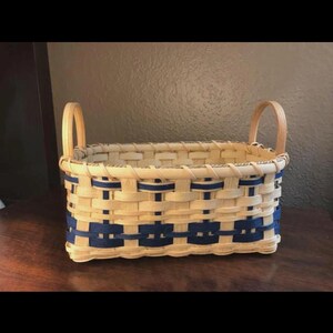May include: A rectangular woven basket featuring light tan natural reed strips interlaced with dark navy blue accent strips. The basket includes two arched handles on the sides and a sturdy base, showcasing a traditional woven pattern throughout.