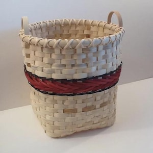 May include: A handwoven basket with a rectangular base and a round top. The basket is made of light-colored woven strips, with a band of red and black woven material around the middle. It has two wooden handles.