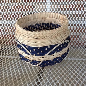 May include: A handwoven basket with a round shape. The basket features alternating bands of natural and navy blue woven material. A decorative white woven element wraps around the navy blue section.