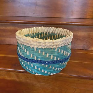 May include: A round woven basket with a light beige rim and a turquoise and blue body. The basket is made of woven material and has a textured appearance. The basket is sitting on a wooden surface.