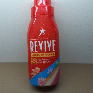 May include: A red bottle of Revive liquid stiffener with a white star and polka dots. The label includes the text "Revive Liquid Stiffener" and "3X Brighter". The bottle has a red cap and a blue and white stripe design with floral accents.