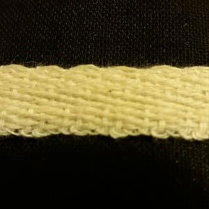 May include: Close-up of a cream-colored woven ribbon against a black background. The ribbon has a textured, ridged appearance, with visible threads and a slightly uneven edge. The weave is tight, creating a sturdy look.