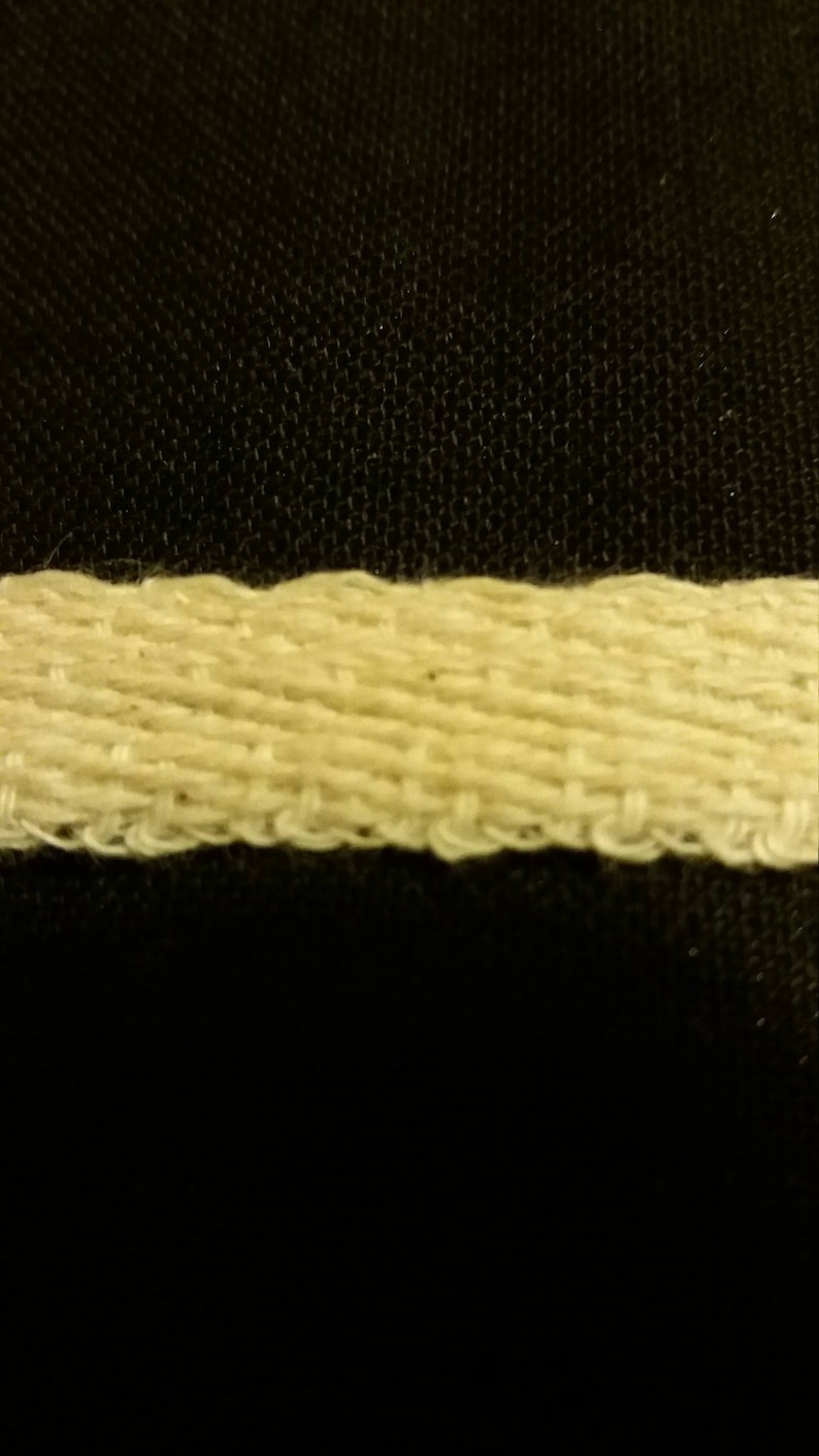May include: Close-up of a cream-colored woven ribbon against a black background. The ribbon has a textured, ridged appearance, with visible threads and a slightly uneven edge. The weave is tight, creating a sturdy look.