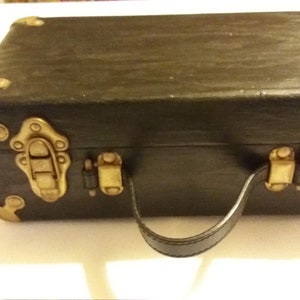 May include: A small black suitcase with gold accents and a leather handle. The suitcase has two latches on each side and a decorative design on the front.