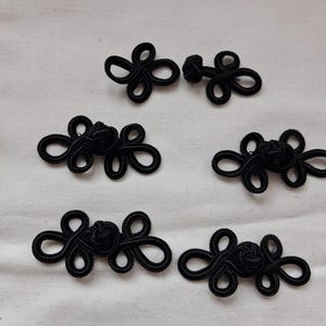 May include: A collection of six black frog closures, also known as Chinese knots, arranged on a white fabric surface. The closures feature intricate looped designs and a central knot, commonly used for fastening garments.