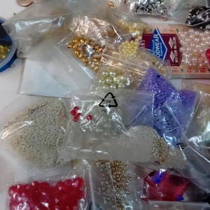 Puede incluir: Surtido de cuentas de colores en bolsas de plástico. Las cuentas son de varias formas, tamaños y colores, incluyendo blanco, rojo, dorado, morado y transparente. Las bolsas están etiquetadas con un símbolo de reciclaje.