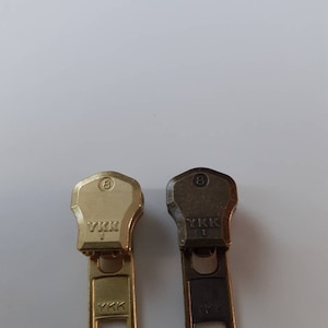 Op de afbeelding: Twee YKK-rits-schuifjes, een gouden en een zwarte, met het nummer 8 bovenaan en de tekst "YKK" aan de voor- en achterkant.