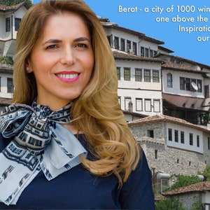 Può includere: Una donna con lunghi capelli biondi indossa una camicia blu e una sciarpa con un motivo di edifici bianchi con finestre. Il testo "Berat - una città di 1000 finestre una sopra l'altra. Ispirazione per la nostra sciarpa Berat" è visibile sullo sfondo.