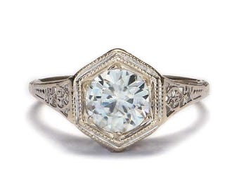 May include: A silver engagement ring with a large, round, clear gemstone set in a hexagonal frame. The ring has intricate floral detailing on the band and around the gemstone setting.