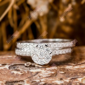 May include: A set of two silver rings with a flower-shaped center stone. The center stone is surrounded by smaller diamonds. The rings are set on a brown wooden surface.