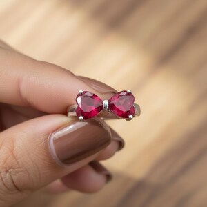 May include: A silver-toned ring featuring two faceted, heart-shaped red gemstones arranged to resemble a bow. The stones are held in place by small metal prongs, creating a symmetrical design on a simple, polished metal band that encircles the finger.
