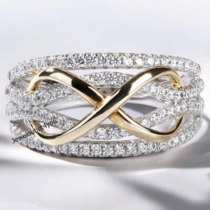 May include: A silver-toned ring with a gold-colored infinity symbol design. The ring features multiple bands encrusted with small, clear gemstones. The infinity symbol is centered and made of polished gold.