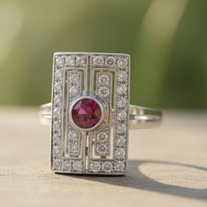 May include: A silver ring with a rectangular face, adorned with small diamonds and a central, round, red gemstone. The ring is set against a blurred green and tan background, showcasing its Art Deco design.