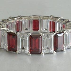 May include: An eternity band ring featuring alternating emerald-cut deep red gemstones and clear white gemstones set in a silver-toned metal band. The stones are arranged in a continuous pattern around the entire circumference of the ring.