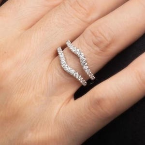 May include: A silver-toned ring guard with a curved design, encrusted with small, sparkling clear stones. The ring is designed to be worn with another ring, creating a layered effect. The ring is shown on a hand.