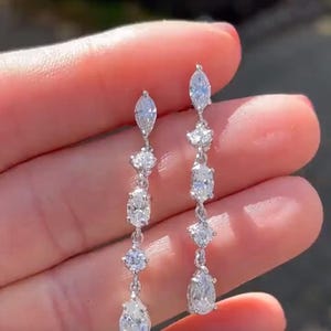 Op de afbeelding: Hangende zilveren oorbellen met meerdere heldere, sprankelende edelstenen. De oorbellen hebben een reeks stenen in verschillende vormen, waaronder markies-, ovale en peervormige slijpsels, verbonden door delicate schakels.