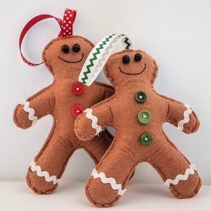Puede incluir: Dos adornos de hombre de jengibre de fieltro con ribetes blancos y botones coloridos. Los hombres de jengibre son marrones y tienen caras sonrientes.