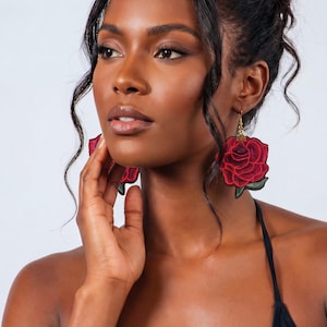 May include: A woman is wearing large, red rose-shaped earrings with green leaves. The earrings are embroidered and have gold-coloured hooks. The woman has dark hair and is wearing a black top.