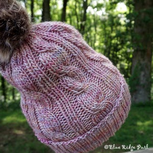 May include: A hand-knitted winter hat in shades of pink, purple, and cream, featuring a cable knit design and a faux fur pom-pom. The hat has a ribbed brim and a textured pattern. The background is a blurred view of a forest.