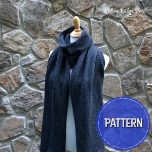 May include: A dark gray knitted scarf with a cowl neck is displayed on a mannequin. The scarf is long and rectangular, with a textured knit pattern. The background is a stone wall. A blue circle with the word "PATTERN" is in the lower right corner.