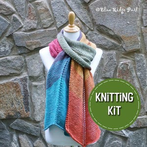 May include: A knitting kit featuring a colourful scarf in shades of teal, orange, purple, and green. The scarf is draped over a wooden mannequin against a stone wall background. The text "KNITTING KIT" is displayed in a green circle.