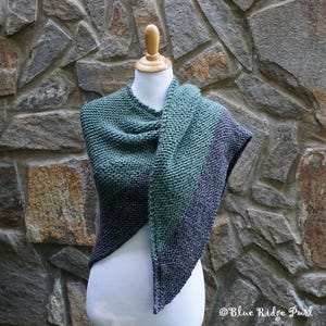 May include: A triangular shawl knitted in a gradient of green and grey yarn. The shawl is draped over a white mannequin against a stone wall background.